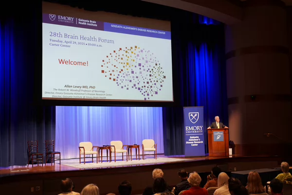 Speaker at a Brain Health Forum at Emory University, standing at a podium beside a large projection screen displaying the event title, date, and welcome message in front of an audience.