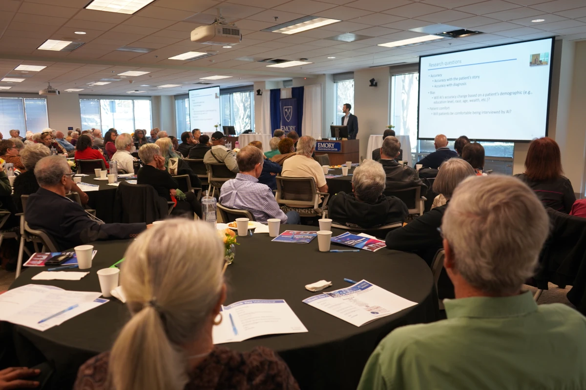 Audience seated at round tables listening to a speaker presenting slides at an Emory Brain Health Center event.